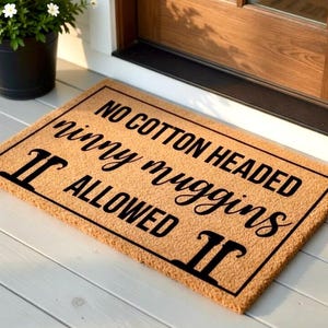 Pode incluir: Um capacho retangular com o texto "NO COTTON HEADED ninny muggins ALLOWED" em preto sobre um fundo bege. O tapete est&aacute; colocado sobre uma superf&iacute;cie de madeira branca perto de uma porta.