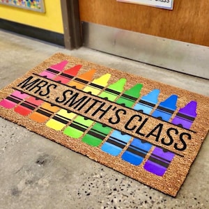 May include: A rectangular doormat with a natural fiber background. The mat features a row of colorful crayon illustrations in a rainbow pattern. The text "MRS. SMITH'S CLASS" is printed in black across the center.