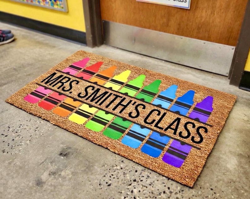 May include: A rectangular doormat with a natural fiber background. The mat features a row of colorful crayon illustrations in a rainbow pattern. The text "MRS. SMITH'S CLASS" is printed in black across the center.