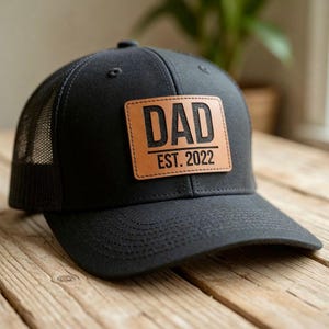 May include: Black trucker hat with a mesh back and a curved brim. A rectangular brown leather patch on the front displays the word "DAD" above "EST. 2022" in black lettering. The hat is sitting on a wooden surface.