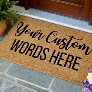 May include: A rectangular doormat made of natural coir fiber. The mat features the text "Your Custom WORDS HERE" in black, stylized lettering. A potted fern is visible in the upper left corner. The mat is placed on a stone surface.
