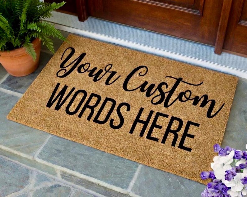 May include: A rectangular doormat made of natural coir fiber. The mat features the text "Your Custom WORDS HERE" in black, stylized lettering. A potted fern is visible in the upper left corner. The mat is placed on a stone surface.
