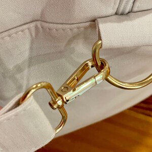 May include: A close-up of a white canvas bag with a gold metal clasp and a gold metal ring. The bag has a zipper closure.