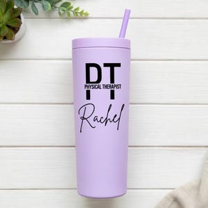 Puede incluir: Un vaso de color lila claro con tapa y pajita a juego. El vaso presenta el texto "DT PHYSICAL THERAPIST Rachel" en negro. El vaso cil&iacute;ndrico parece ser de metal o material similar.