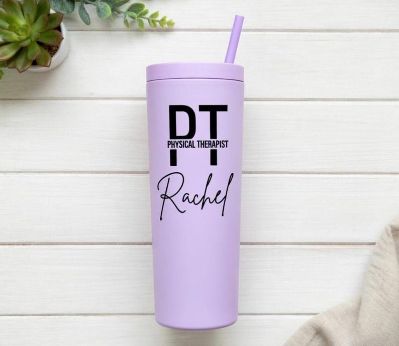 Puede incluir: Un vaso de color lila claro con tapa y pajita a juego. El vaso presenta el texto "DT PHYSICAL THERAPIST Rachel" en negro. El vaso cil&iacute;ndrico parece ser de metal o material similar.