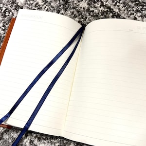 May include: A brown leather-bound journal with two blue ribbon bookmarks. The journal has lined pages and a space for the date at the top of each page.