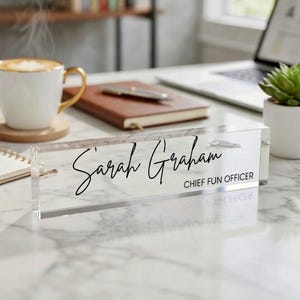 May include: A clear acrylic desk nameplate with the name "Sarah Graham" in elegant script and the title "CHIEF FUN OFFICER" in a smaller, block font. The nameplate is on a marble-patterned surface.