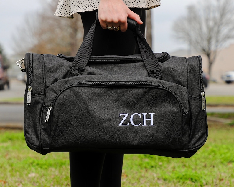 May include: A black duffel bag with a white monogram "ZCH" embroidered on the front. The bag has two zippered compartments and a top handle.
