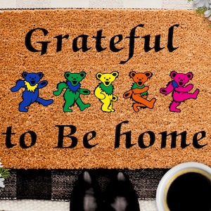 May include: A brown coir doormat with the text "Grateful to Be Home" in black lettering. Five colorful dancing bears are arranged in a row above the text.