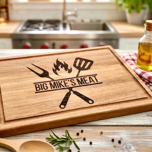 Puede incluir: Tabla de cortar de madera con un dise&ntilde;o tallado que presenta un tenedor, una esp&aacute;tula, llamas y el texto "BIG MIKE'S MEAT". La tabla tiene un surco para el jugo y est&aacute; rodeada por un marco de madera. Una peque&ntilde;a botella de vidrio de aceite y un pa&ntilde;o a cuadros rojo y blanco est&aacute;n en el fondo.