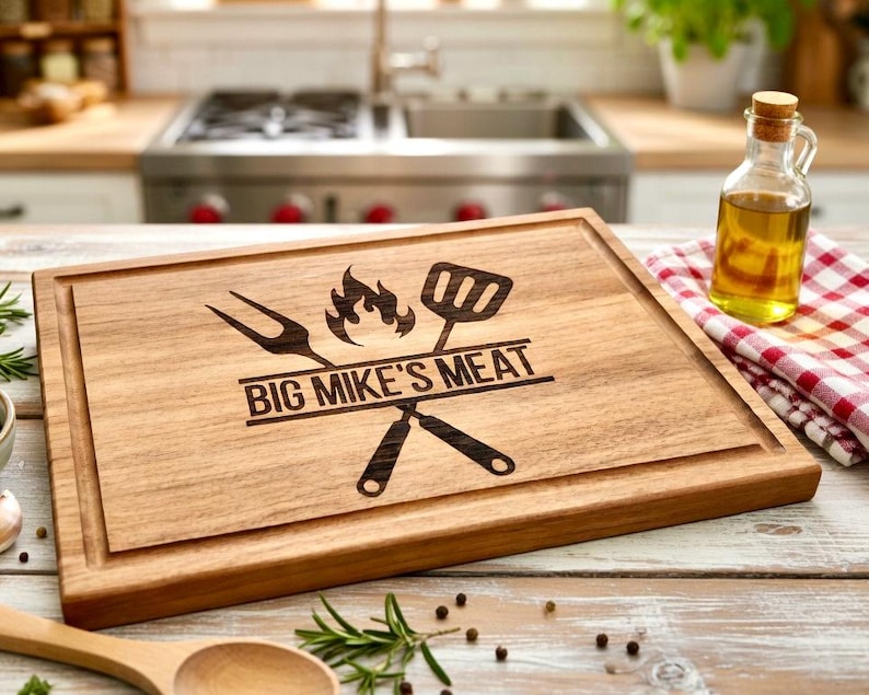 Puede incluir: Tabla de cortar de madera con un dise&ntilde;o tallado que presenta un tenedor, una esp&aacute;tula, llamas y el texto "BIG MIKE'S MEAT". La tabla tiene un surco para el jugo y est&aacute; rodeada por un marco de madera. Una peque&ntilde;a botella de vidrio de aceite y un pa&ntilde;o a cuadros rojo y blanco est&aacute;n en el fondo.