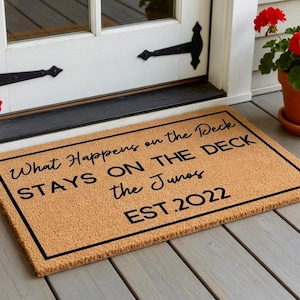 May include: A rectangular doormat with a black border and text that reads "What Happens on the Deck STAYS ON THE DECK the Junos EST.2022". The mat is made of a natural fiber material and is placed in front of a white door.
