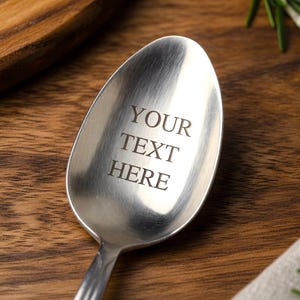 May include: A stainless steel spoon with the engraved text "YOUR TEXT HERE". The spoon is set against a wooden surface, with sprigs of rosemary in the background. The spoon's design is simple and elegant.
