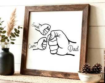 May include: A framed wall art print featuring a black and white illustration of three fists bumping. The names "Sarah", "Cam", and "Dad" are written above or near each fist. The frame is made of dark brown wood.