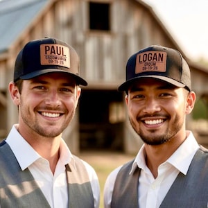 May include: Two black trucker caps with leather patches. One cap reads "PAUL GROOMSMAN 09-15-26" and the other reads "LOGAN GROOM 09-15-26". The caps are worn by two men, both smiling. The background is a wooden barn.