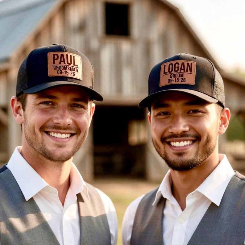 May include: Two black trucker caps with leather patches. One cap reads "PAUL GROOMSMAN 09-15-26" and the other reads "LOGAN GROOM 09-15-26". The caps are worn by two men, both smiling. The background is a wooden barn.