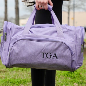 Pu&ograve; includere: Una borsa da viaggio di colore lavanda con chiusura a zip e manico superiore. La borsa ha un monogramma "TGA" ricamato sul davanti.