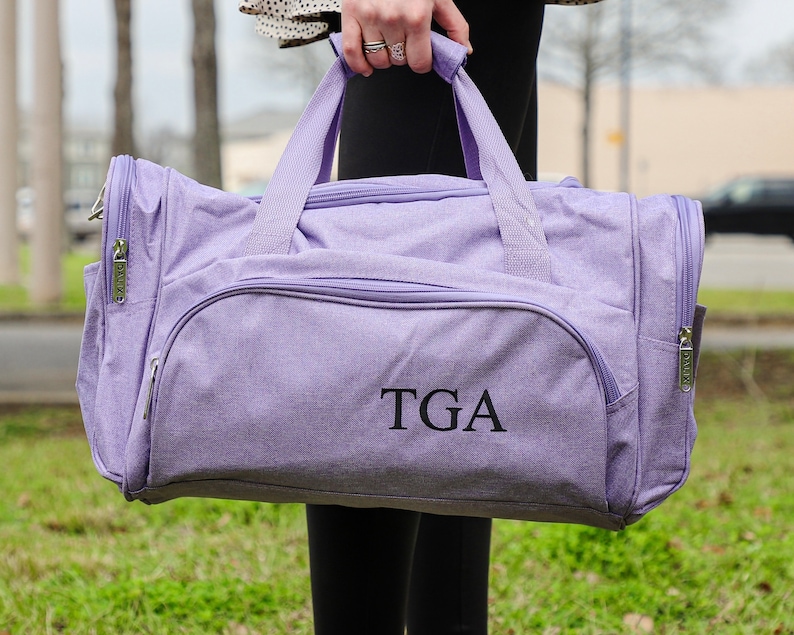Pu&ograve; includere: Una borsa da viaggio di colore lavanda con chiusura a zip e manico superiore. La borsa ha un monogramma "TGA" ricamato sul davanti.