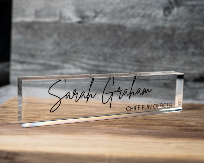 May include: A clear acrylic desk nameplate displaying the name "Sarah Graham" in a cursive font. The title "CHIEF FUN OFFICER" is printed below the name. The nameplate is positioned on a wooden surface, with a grey background.