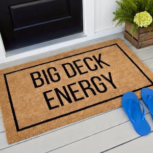 May include: A rectangular doormat with the words "BIG DECK ENERGY" in black block letters. The mat is brown with a black border and is placed in front of a black door. Two blue flip-flops are next to the mat.