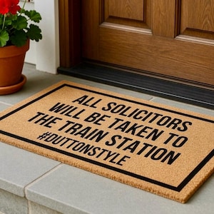 May include: Rectangular doormat with a light brown background and a black border. The text "ALL SOLICITORS WILL BE TAKEN TO THE TRAIN STATION #DUTTONSTYLE" is printed in black across the mat. The doormat is placed in front of a wooden door.