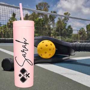 K&ouml;nnte beinhalten: Ein rosa, personalisierter Becher mit Strohhalm, mit dem Namen "Sarah" und einer schwarz-wei&szlig;en Grafik von zwei Pickleball-Schl&auml;gern und einem Ball. Der Becher steht auf einem Pickleball-Platz mit einem Pickleball-Schl&auml;ger und einem Ball im Hintergrund.