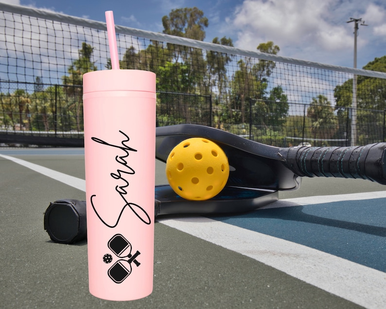 K&ouml;nnte beinhalten: Ein rosa, personalisierter Becher mit Strohhalm, mit dem Namen "Sarah" und einer schwarz-wei&szlig;en Grafik von zwei Pickleball-Schl&auml;gern und einem Ball. Der Becher steht auf einem Pickleball-Platz mit einem Pickleball-Schl&auml;ger und einem Ball im Hintergrund.