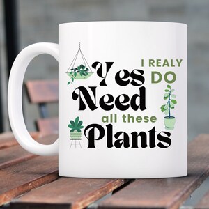 K&ouml;nnte beinhalten: Wei&szlig;e Keramiktasse mit einem schwarzen und gr&uuml;nen Design. Die Tasse zeigt den Text "I really do need all these plants" mit Illustrationen von Topfpflanzen.