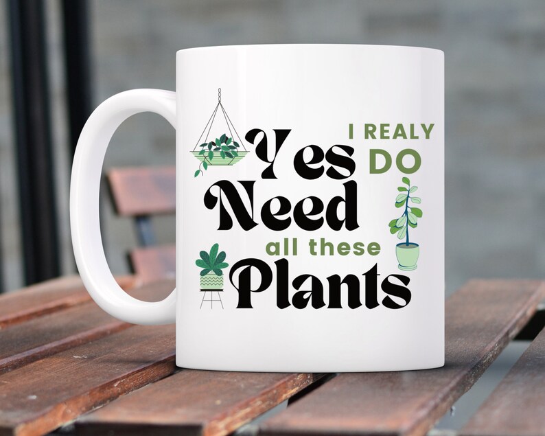 K&ouml;nnte beinhalten: Wei&szlig;e Keramiktasse mit einem schwarzen und gr&uuml;nen Design. Die Tasse zeigt den Text "I really do need all these plants" mit Illustrationen von Topfpflanzen.