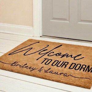 Op de afbeelding: Een rechthoekige deurmat van natuurlijke vezels met de zwarte tekst "Welcome TO OUR DORM" en de namen "Britney & Laura" eronder. De mat ligt voor een witte deur.