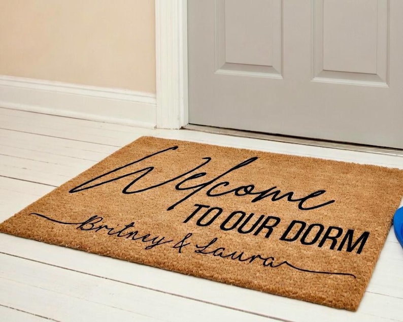 Op de afbeelding: Een rechthoekige deurmat van natuurlijke vezels met de zwarte tekst "Welcome TO OUR DORM" en de namen "Britney & Laura" eronder. De mat ligt voor een witte deur.