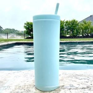 May include: A light blue stainless steel tumbler with a straw. The tumbler is sitting on a concrete surface next to a swimming pool.