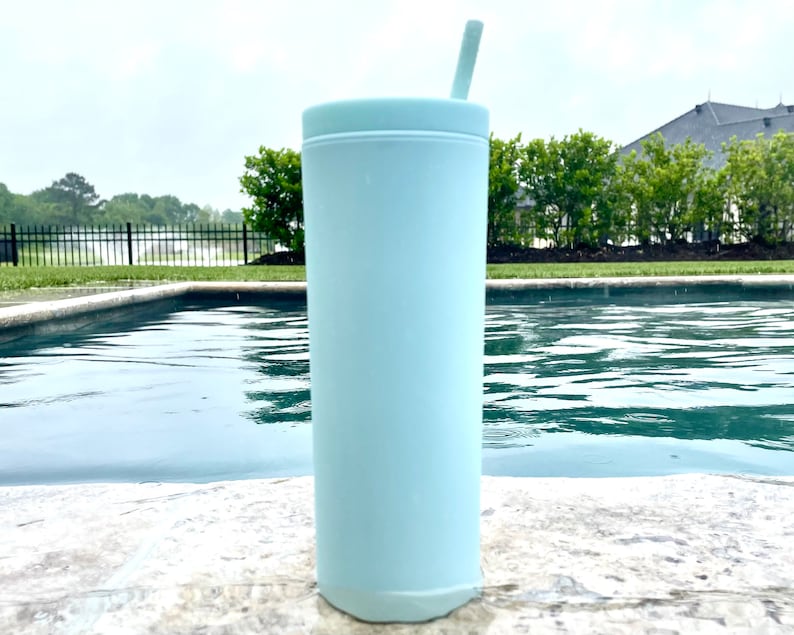 May include: A light blue stainless steel tumbler with a straw. The tumbler is sitting on a concrete surface next to a swimming pool.