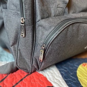 May include: Close-up of a gray duffel bag with two zipper pulls that have the word "DALIX" printed on them. The bag has a black zipper and a black pocket.