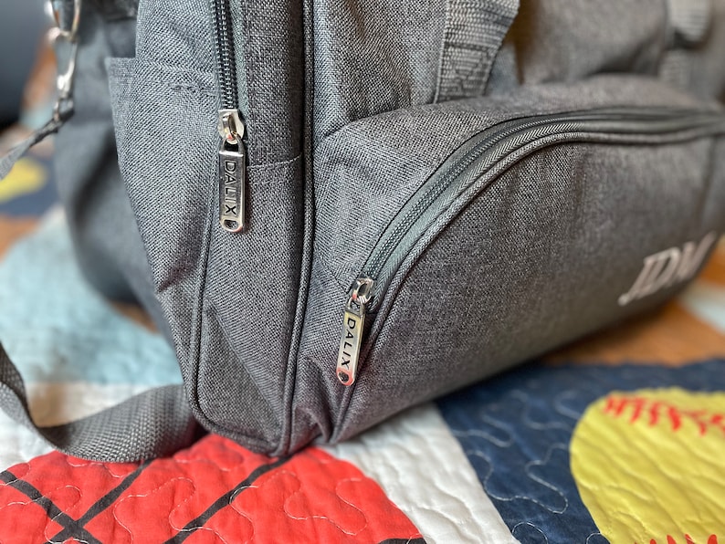 May include: Close-up of a gray duffel bag with two zipper pulls that have the word "DALIX" printed on them. The bag has a black zipper and a black pocket.