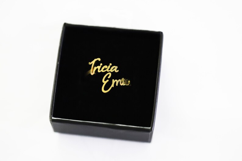 May include: A gold ring with the names "Tricia" and "Emma" engraved in cursive script. The ring is displayed in a black velvet box.