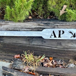 Puede incluir: Una pala de metal con las iniciales "AP" y una silueta de cangrejo de r&iacute;o. La pala est&aacute; pintada de gris claro y descansa sobre un tronco de madera.