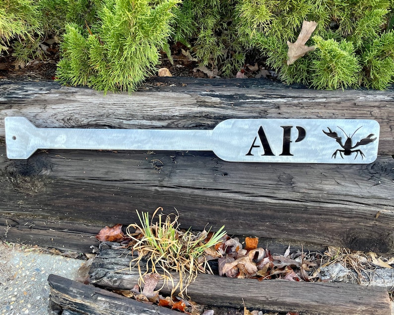 Puede incluir: Una pala de metal con las iniciales "AP" y una silueta de cangrejo de r&iacute;o. La pala est&aacute; pintada de gris claro y descansa sobre un tronco de madera.