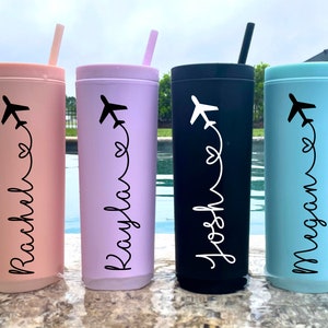 May include: Four personalized travel tumblers with a plane and heart design. The tumblers are pink, purple, black, and blue. The names on the tumblers are Rachel, Kayla, Josh, and Megan.