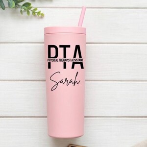Puede incluir: Un vaso rosa con tapa y pajita a juego. El vaso presenta el texto "PTA PHYSICAL THERAPIST ASSISTANT" en letras negras, con el nombre "Sarah" en escritura cursiva debajo.