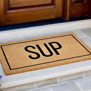 May include: A rectangular tan coir doormat featuring a black rectangular border and the word SUP printed in large black capital letters in the center. The mat has a coarse, fibrous texture and is positioned horizontally on a flat surface.
