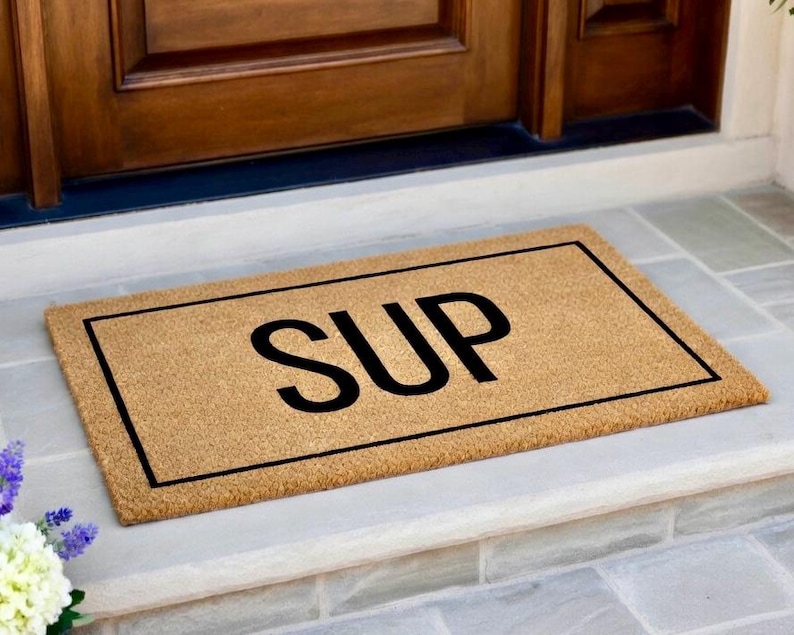 May include: A rectangular tan coir doormat featuring a black rectangular border and the word SUP printed in large black capital letters in the center. The mat has a coarse, fibrous texture and is positioned horizontally on a flat surface.