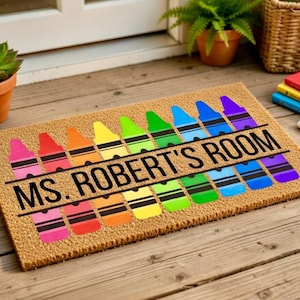 May include: A rectangular doormat made of natural fibers, featuring a colorful crayon design. The crayons are arranged in a row, with the text "MS. ROBERT'S ROOM" in black. The mat is placed on a wooden surface.