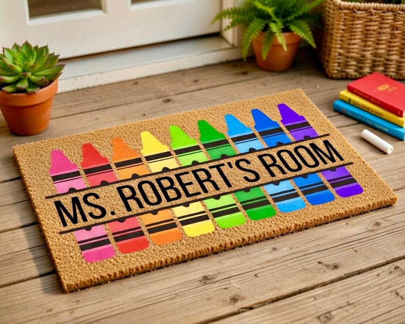 May include: A rectangular doormat made of natural fibers, featuring a colorful crayon design. The crayons are arranged in a row, with the text "MS. ROBERT'S ROOM" in black. The mat is placed on a wooden surface.