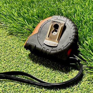 May include: A black and orange tape measure with a black strap, lying on a green artificial grass surface.