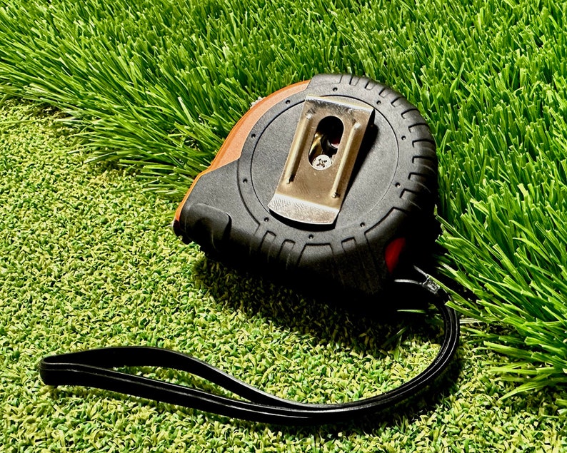 May include: A black and orange tape measure with a black strap, lying on a green artificial grass surface.