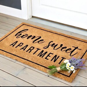 May include: A rectangular doormat made of natural fibers with the words "home sweet APARTMENT" in black script. A small bouquet of lavender and daisies rests on the mat. The mat is bordered with a black frame.