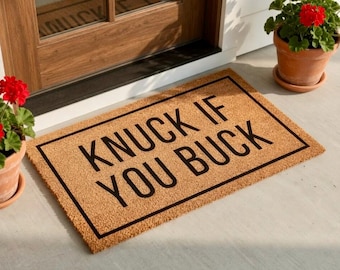 May include: A rectangular doormat made of natural coir fiber, featuring the phrase "KNUCK IF YOU BUCK" in bold, black capital letters. The mat is framed with a black border and sits on a light-colored surface near a wooden door.