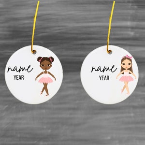May include: Two white ceramic ornaments with a ballerina illustration. Each ornament has the text "name" and "year" on it. One ballerina has brown hair and a pink tutu, the other has brown skin, brown hair, and a pink tutu.