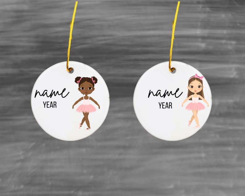 May include: Two white ceramic ornaments with a ballerina illustration. Each ornament has the text "name" and "year" on it. One ballerina has brown hair and a pink tutu, the other has brown skin, brown hair, and a pink tutu.